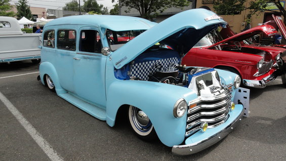 Light blue custom Chevrolet Suburban with hood open, showing engine bay with checkerboard pattern. Red car behind it