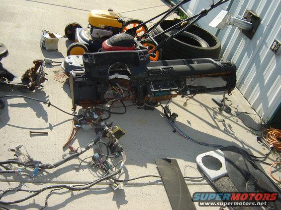 Disassembled car dashboard with extensive wiring on concrete, next to lawnmowers and tires