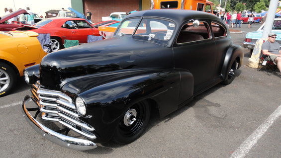 Black hot rod coupe with chrome grille and bumper, parked at a car show. Other cars visible in background