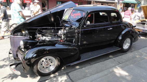 black coupe hot rod with hood open, showing engine and chrome grille. Another car is visible to the right