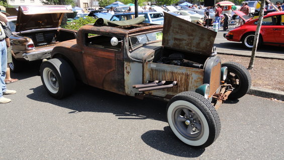 rusted hot rod with open hood and exhaust pipes, parked next to a brown car with open trunk