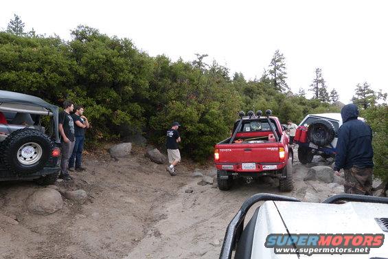 Several off-road vehicles, including a red Toyota pickup, are on a rocky dirt trail with trees in the background