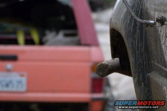 Muddy exhaust pipe on a vehicle, with a pink SUV in the background. License plate visible on the SUV