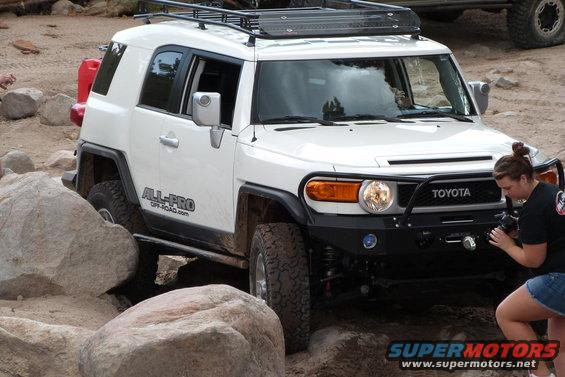 White Toyota FJ Cruiser navigating rocks on a trail, with a person filming in the foreground