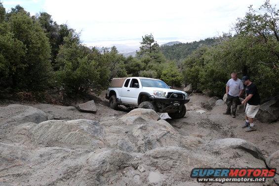 White Toyota Tacoma pickup truck with a tan soft topper is navigating a rocky, off-road trail. Two men stand nearby watching