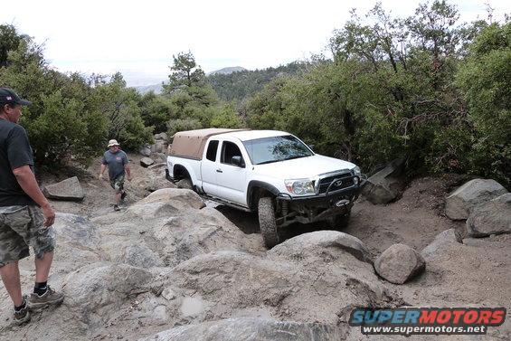 White Toyota pickup truck with a tan soft top navigating a rocky trail, front bumper modified