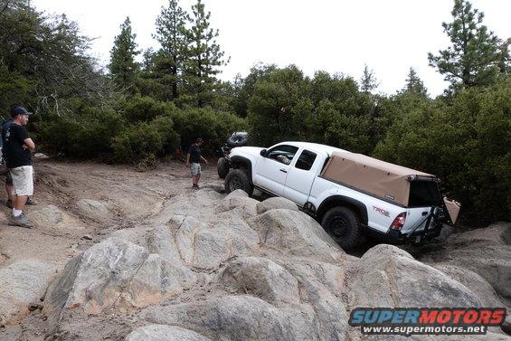 White Toyota Tacoma pickup truck with tan camper shell navigating a rocky trail