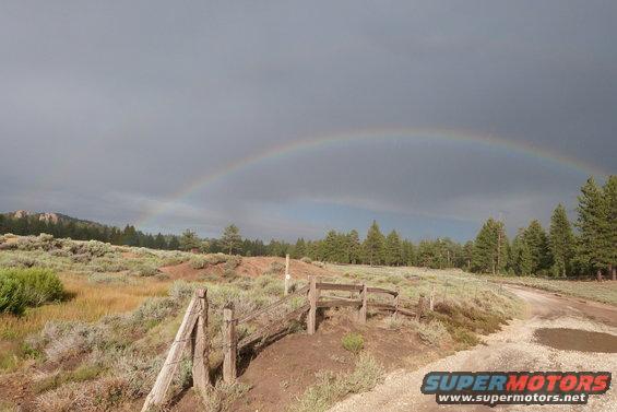 Rural landscape with a wooden fence in the foreground and a rainbow arching across a cloudy sky
