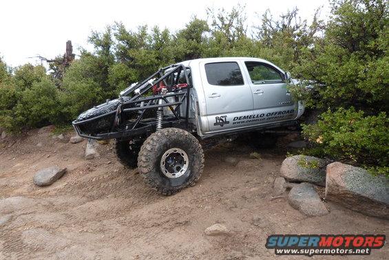 Silver Toyota Tacoma with custom roll cage and large off-road tires navigating rocky terrain