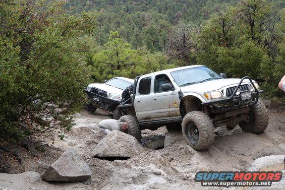 Two modified trucks on a rocky trail, one with a person underneath