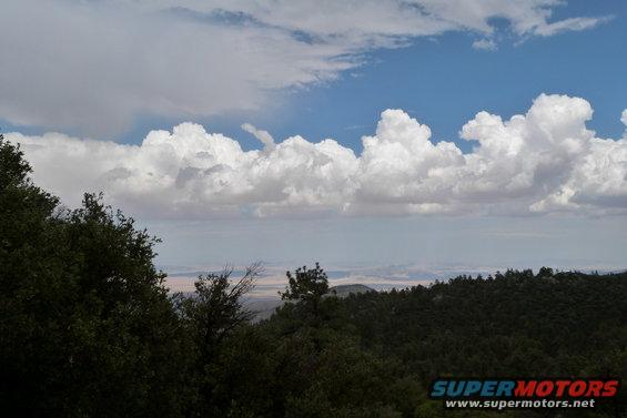 Overcast sky with large white clouds above a dark green forested mountain range and a distant hazy valley