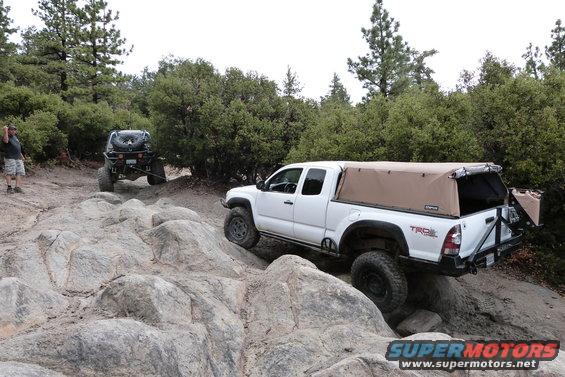 White Toyota Tacoma with a tan soft topper driving over rocks on a trail. Another off-road vehicle is behind it