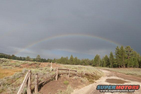 Dirt road with puddles, wooden fence, and a rainbow over a treeline
