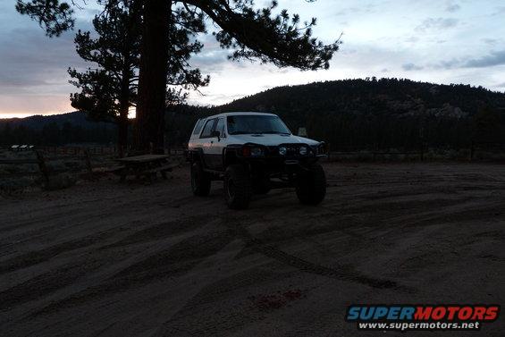 White SUV with off-road tires and brush guard parked at dusk near trees and hills