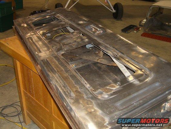 Car door with internal mechanisms exposed, resting on a wooden surface. Another vehicle is partially visible in the background