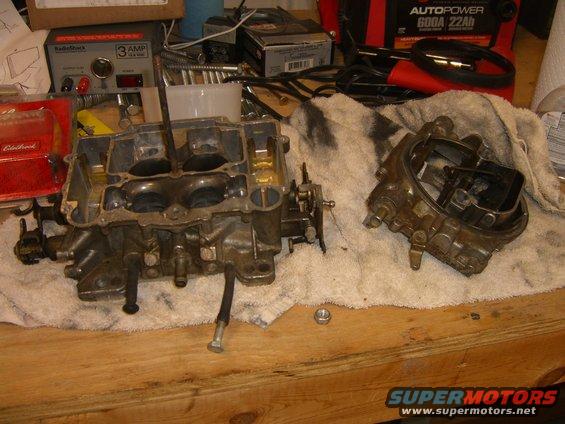 Two carburetors disassembled on a workbench with tools and a battery charger in the background