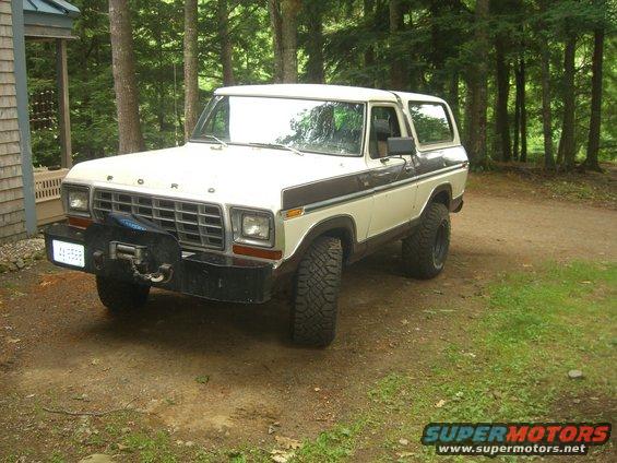 White and brown Ford Bronco with a winch on the front bumper parked on a dirt road near a house and trees