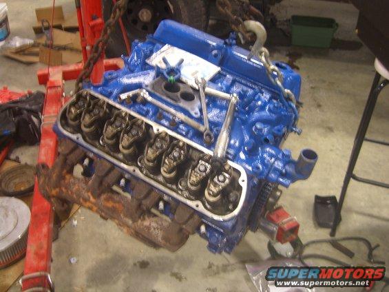 Blue V8 engine with valve covers off, being lifted by a red engine hoist. Wrench on top