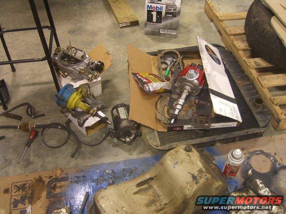 Car parts including a carburetor, distributor, and oil pan are scattered on a concrete floor