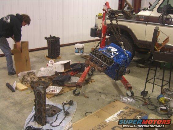 Engine on a hoist, with another engine block on the floor. A person is handling a box. A truck is in the background