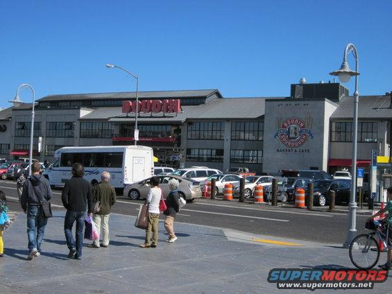 People walking in front of Boudin Bakery with cars and a bus parked nearby