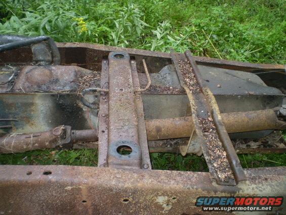 rusted vehicle frame showing fuel tank and driveshaft, surrounded by grass