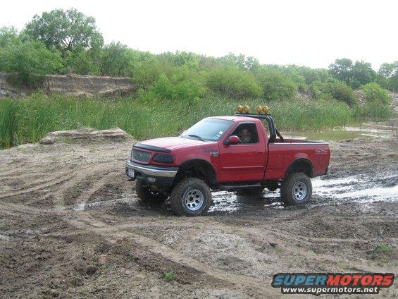 Red lifted Ford F-150 pickup truck driving through mud with a roll bar and yellow lights