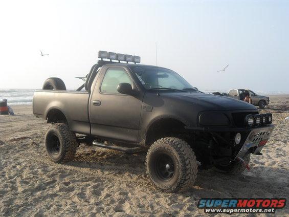 Modified black pickup truck with off-road tires and light bar on a sandy beach. Another truck is visible in the background