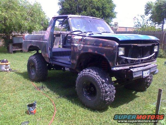 Rusty Ford Bronco with lifted suspension and large tires, doors removed, blue hood