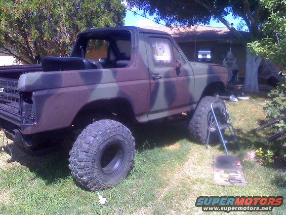 Camouflaged Ford Bronco with oversized tires, missing front grille, and a spare tire on a stand