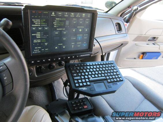 Interior view of a police car showing a large computer screen, keyboard, and control panel. The screen displays various radio and patrol functions. The dashboard and seats are visible