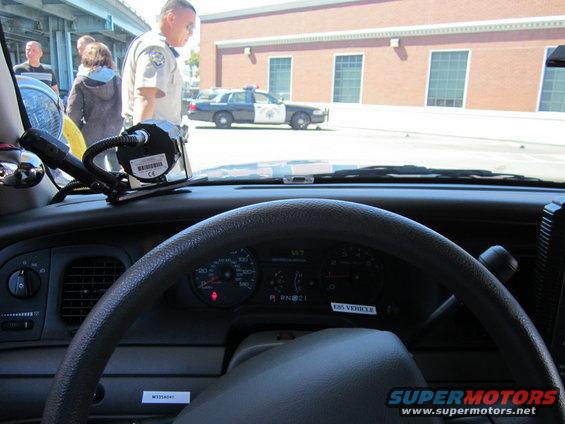 Police car interior dashboard with steering wheel, gauges, and radio equipment. A police car is visible outside