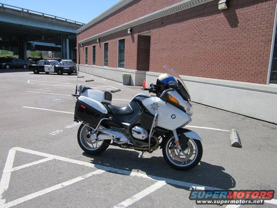 White police motorcycle parked in a lot, with police cars in the background