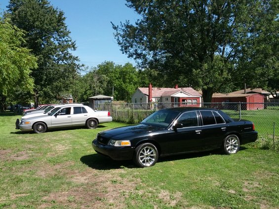 Black Ford Crown Victoria with chrome wheels parked on grass, 3/4 front view. Another silver Crown Victoria is parked behind it