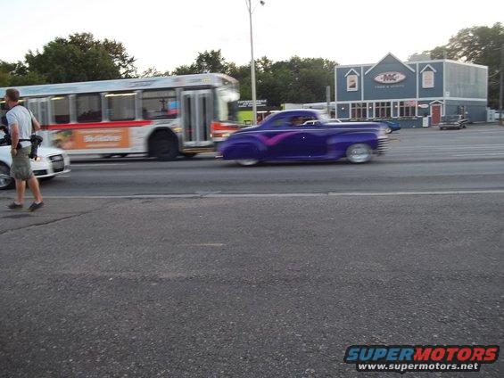 Purple customized coupe with pink racing stripes driving on a street next to a bus