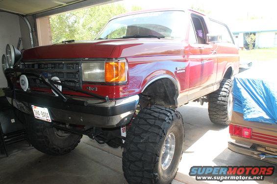 Red lifted Ford Bronco with large off-road tires parked in a garage. Has extra lights on the front
