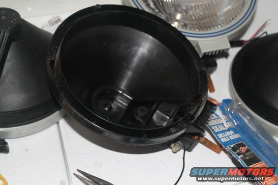 Close-up of a black plastic headlight housing with a headlight assembly and another housing nearby. A tube of black sealant is also visible