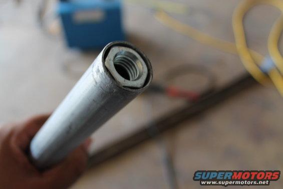 Close-up of a metal pipe with a nut inside, held by a hand. A blue box and yellow cable are blurred in the background