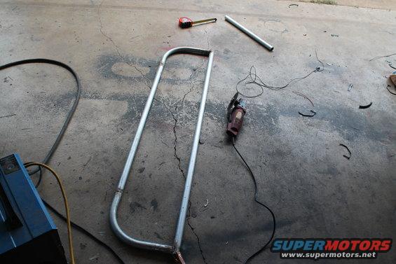 Metal tubing bent into a U-shape, with a grinder, tape measure, and welding equipment on a concrete floor