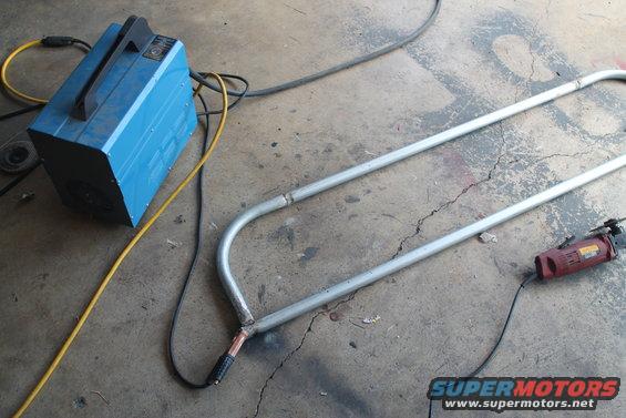 Blue welding machine with yellow and black cables next to bent metal tubing and a red grinder
