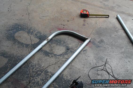 Metal tubing bent into a U-shape lies on a concrete floor next to a tape measure and wires