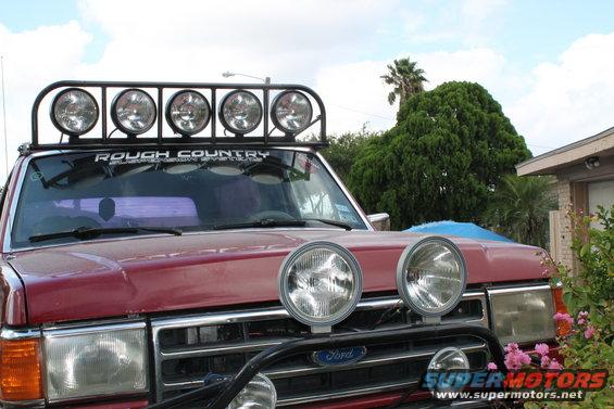 Red Ford Bronco with a roof-mounted light bar and front-mounted fog lights. "Rough Country" sticker on windshield