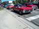 1970 Ford Ranchero - A line of cars, including a red convertible and a red pickup truck, are driving down a street on a sunny day.