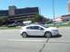 1970 Ford Ranchero - A white sedan is captured in motion on a sunny day, with a large building and other vehicles visible.