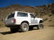 1996 Ford Bronco - This modified SUV is shown on a dusty trail with rocky hills under a clear blue sky.
