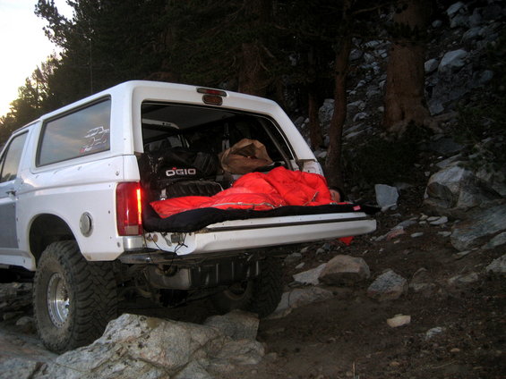 White Ford Bronco with open tailgate, red sleeping bag, and camping gear inside, parked on rocky terrain