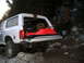 1996 Ford Bronco - A white Ford Bronco is parked on a rocky trail with its tailgate open, revealing camping gear and a sleeping bag.