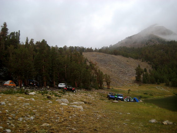 Campsite with tents and several off-road vehicles parked on a grassy slope near trees and a rocky hill
