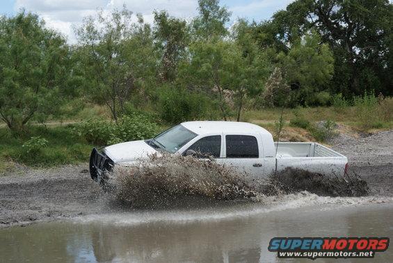 White pickup truck splashing through a muddy puddle with trees in the background