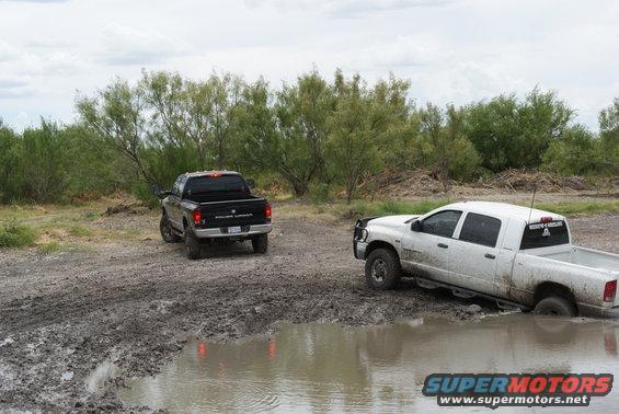 White pickup truck stuck in mud puddle, black pickup truck nearby